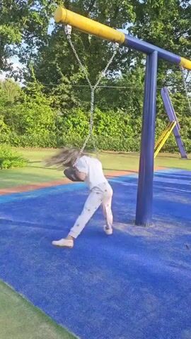 A young girl with blonde hair is swinging on a swing set at a playground. She is wearing a light-colored shirt and light-colored pants. Her hair is flying around her as she swings. The swing set has a yellow top bar and blue poles. The ground beneath the swing set is covered in blue rubber matting. In the background, there are trees and green grass. The lighting suggests it is daytime.