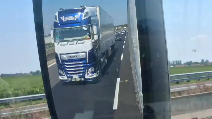 The video is a first-person perspective shot from inside a vehicle, looking into a side mirror. The mirror reflects a blue and white DAF semi-truck with a white trailer driving on a highway. The truck appears to be in the lane to the right of the vehicle from which the video is being filmed. The highway is multi-lane with white dashed lines separating the lanes. In the distance, traffic is visible, suggesting a congestion or slowdown. The environment outside the mirror shows a bright, sunny day with green fields and trees bordering the highway. Another car is visible in the mirror on the left side of the frame, appearing to be in the lane to the left of the truck. The reflection in the mirror is the primary focus of the video.