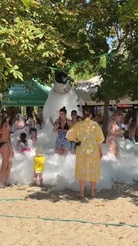 The video captures a lively outdoor scene, likely at a beach or poolside, filled with people enjoying a foam party. In the center, a large, mound of white foam rises, resembling a giant snowman. People, including adults and children, are immersed in the foam, with some appearing to play and interact. A young child in a yellow shirt stands prominently in the foreground, facing the foam. Several adults are visible around the foam mound. The environment is sunny, with lush green trees providing shade overhead. Beach umbrellas and some structures are visible in the background, suggesting a recreational establishment. The overall atmosphere appears festive and energetic.