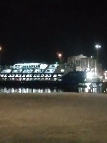 The video shows a lit-up ship docked in a harbor at night. The ship is oriented vertically, with the harbor to the right and a dark, undefined area to the left. The ship has several decks and rows of lit windows.  There are lights on the upper deck and possibly a small tower or structure. The water reflects the lights from the ship and surrounding area.  The perspective appears to be from ground level, slightly elevated, looking out at the ship.