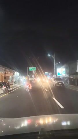 A first-person perspective video taken from inside a vehicle at night. The vehicle is on a multi-lane road with traffic. Streetlights and vehicle headlights illuminate the scene, creating lens flare effects. Several motorcycles are visible in the foreground and middle ground, some with illuminated taillights. Cars and a bus are also present in the traffic. Road signs with green backgrounds and white text are visible, indicating directions. Buildings line the right side of the road, some with illuminated signs. The overall environment suggests an urban or suburban setting during nighttime hours.