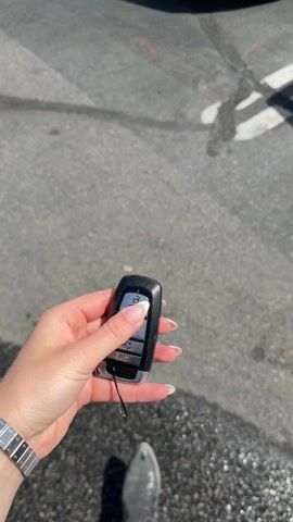The video shows a person holding a car key fob in their hand, with a silver convertible Mustang in the background. The car is parked in a parking lot on a sunny day. The person presses a button on the fob, and the car's tail lights flash. The person is wearing a silver watch and has white nail polish. Other cars are visible in the background, including a white pickup truck. The license plate on the Mustang reads "BRD 1668".