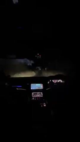The video shows the dashboard and front view from inside a car driving at night. The road is dark, illuminated by the car's headlights. There are trees and foliage visible on either side of the road. The car's dashboard is visible with a lit-up screen displaying navigation. A person is seen driving the car. Music plays in the background.