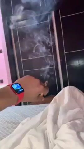 The video shows a person lying in bed, under a white duvet. They are holding a lit, rolled cigarette in their fingers. Smoke is visible, rising from the cigarette. The person has a red Apple Watch on their wrist. The background shows a black tiled wall with thin white grid lines. The watch screen displays the time as 13:20.