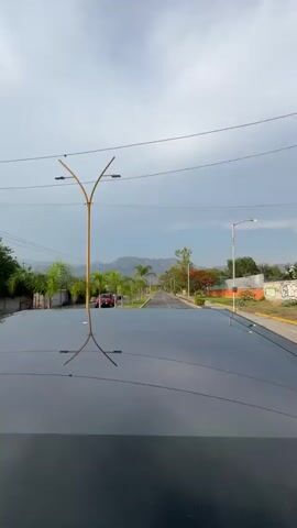 The video shows a view from the roof of a moving vehicle driving down a road. The sky is overcast with a mix of white clouds and a hint of blue. There are palm trees and other trees lining the road, with some trees having red flowers. Street lights are visible along the road. In the distance, a mountain range can be seen. Another car, which is red, is traveling in the opposite direction on the same road.