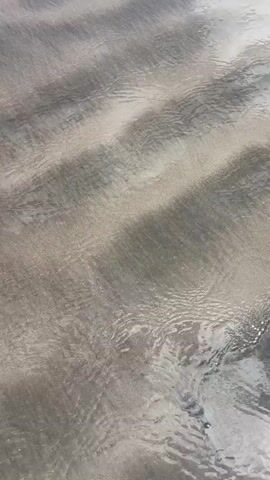 A close-up, vertically-oriented shot shows a sandy beach surface covered by a thin layer of water. The water creates a rippling effect over the sand, revealing variations in color from light tan to darker gray. A pair of feet wearing pink flip-flops is visible in the lower left corner of the frame. The scene is bright, likely taken during the daytime, and conveys a sense of tranquility.