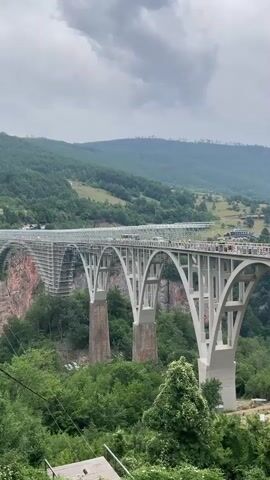 This media file captures a panoramic view of the iconic Đurđevića Tara Bridge in Montenegro. The bridge, a magnificent concrete arch structure, spans a dramatic gorge carved by the Tara River. In the foreground, lush green trees and ferns carpet the steep slopes of the gorge, creating a vibrant natural setting. The bridge itself is the central subject, with its impressive arches and solid concrete piers dominating the landscape.

Several people are visible on the bridge's walkway, some appearing to walk or gather in small groups, enjoying the breathtaking views. A few vehicles, including a silver car, are also crossing the bridge, indicating it's open to traffic. The surrounding landscape is characterized by rolling, densely forested mountains that stretch into the distance, their peaks partially obscured by a cloudy sky. The time of day appears to be daytime, likely late morning or afternoon, given the diffused light. The weather is overcast, with dramatic clouds contributing to the atmospheric mood of the scene. The overall impression is one of awe-inspiring natural beauty and impressive human engineering. No visible text is present in the media file.