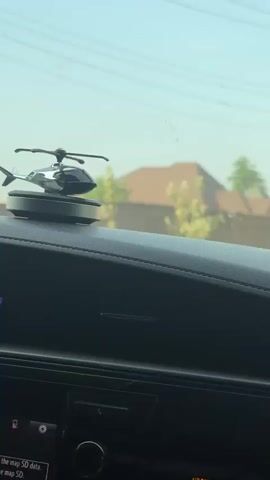The video shows a dashboard of a car, with a decorative miniature helicopter placed on top. The helicopter has black and silver elements, and its rotor blades are spinning. The time displayed on the car's digital clock reads 5:20. Outside the car window, a residential area with houses and greenery is visible under a slightly hazy sky.