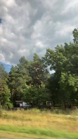 A view from a moving vehicle captures a lush green landscape. Dense trees, primarily tall pines and deciduous trees, line the right side of the frame. A dirt road is visible, and on the left side of the road, a small, white van is parked next to a small structure, possibly a shelter or shed. The sky above is partly cloudy, with dramatic white clouds against patches of blue. The motion of the vehicle creates a slight blur in the foreground, emphasizing the passing scenery. A signpost with a yellow sign and red trim is visible on the left side of the road near the trees. Another red sign is visible further down the road. The overall impression is a serene drive through a wooded area.