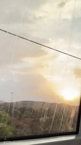 A sunset is viewed through a window, likely from a vehicle. The sun appears as a bright, round orb on the right side of the frame, casting a golden glow across the sky. The sky is filled with scattered clouds, ranging in color from white to yellow and grey, creating a dramatic backdrop. In the foreground, a dark silhouette of hills and trees obscures the lower part of the landscape. A thin, dark wire runs horizontally across the top portion of the window. The window itself appears dirty, with streaks and smudges obscuring the view. The overall impression is one of a scenic journey as the day concludes.