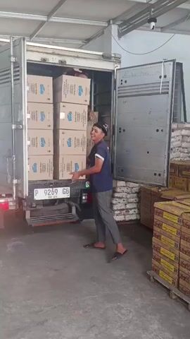 A man in a dark t-shirt, dark shorts, and sandals is standing by the back of a truck, holding a stack of large cardboard boxes. The truck is open, and more boxes are visible inside. The man is smiling and looking towards the camera. To the right of the truck, there are stacks of pallets with more boxes. The floor is concrete, and the lighting suggests an indoor or covered space, possibly a warehouse. The man then picks up the stack of boxes and walks away from the truck, with the camera following him. He is carrying the boxes precariously, with the stack almost as tall as him. He walks past more stacks of boxes on pallets and enters another section of the building. The video is shot from a slightly low angle, looking up at the man and the truck.
