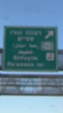 The video shows a road sign with Hebrew and Arabic text, indicating an exit. The sign is yellow with black lettering and an arrow pointing to the right. The sign is located on an overpass, and there are cars and traffic visible in the background. The sky is clear and blue.