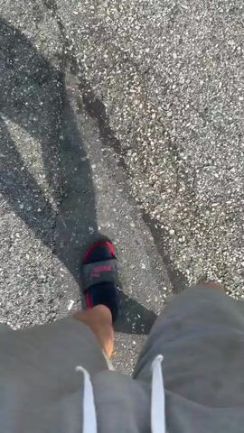 This video shows a first-person perspective of someone walking on an asphalt surface. The person is wearing grey shorts and black socks with red and black slides. The sun is casting a shadow from the person onto the ground. The asphalt appears to be a mix of grey and black aggregate. There is a wet patch on the asphalt to the left of the frame. The video is shot from a downward angle, focusing on the person's legs and feet as they walk.