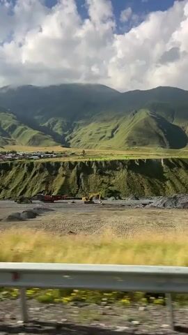The video pans across a construction site in a mountainous, rural area. Several white dump trucks are parked in a dusty lot. In the background, a yellow excavator is visible, along with piles of gravel and two tall, red industrial silos. The construction site is situated at the base of a steep, green, terraced hillside. Beyond the hillside, a small village with scattered houses sits on a wide, flat plain. In the far distance, large, lush green mountains rise, their peaks partially obscured by dramatic, white, cumulus clouds against a bright blue sky. The foreground shows a blur of green foliage and a gray metal guardrail, indicating the viewpoint is from a moving vehicle. The overall scene depicts an active construction or quarrying operation in a scenic, natural landscape.