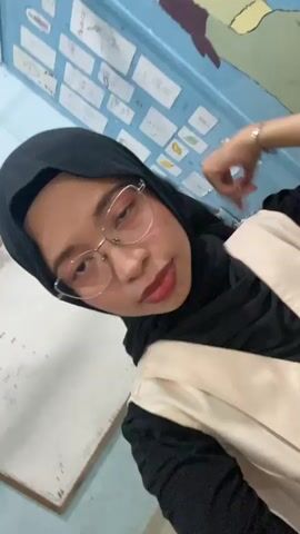 The media file is a selfie-style video clip featuring a young woman wearing a black hijab and glasses. She is dressed in a black top underneath a cream-colored vest. The background appears to be a classroom or educational setting, with a whiteboard on the left and a wall covered in drawings or student work on the right. The drawings on the wall are varied, some appearing to be illustrations and others possibly diagrams or charts. The lighting suggests it is daytime.

The woman is actively engaging with the camera. She is seen touching her forehead and then making a peace sign with her fingers. Her facial expression is playful and energetic, with her tongue slightly sticking out at one point. The overall mood is lighthearted and youthful. There is no discernible text clearly visible in the media file that provides specific information. Given the provided location context, it is likely this scene takes place in Tanjung Gading, Indonesia.