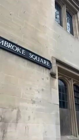 The camera pans up the side of a multi-story light-colored stone building. The building has multiple arched windows with black frames. A street sign reading “BROKE SQUARE” is attached to the building’s wall.  A black gas lamp is mounted on the side of the building. In the distance, a person dressed in black with a white face covering stands in front of a building entrance. The sky is overcast.