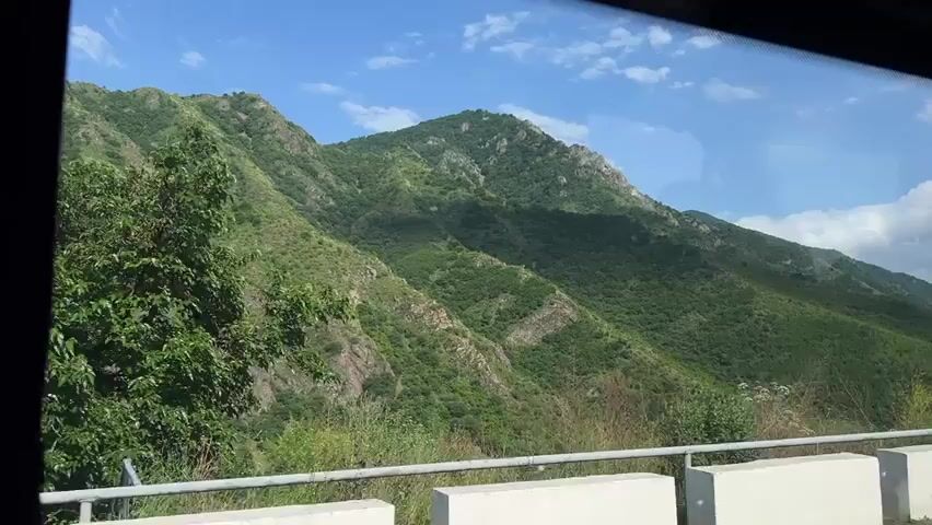 This media file is a video showcasing a scenic mountain landscape, viewed from the perspective of someone inside a vehicle. The foreground features a white concrete barrier and a metal railing, suggesting the view is from a road. Beyond this, a lush, green mountainside dominates the scene, with varied terrain and rocky outcrops visible. The mountains are covered in dense vegetation, appearing to be a mix of trees and scrub. The sky is a clear blue with scattered white clouds, indicating fair weather. The sunlight casts shadows across the mountains, suggesting it is daytime, likely late morning or early afternoon. No people are visible in the frame. The setting appears to be mountainous and rural. There is no visible text within the media file. The location context provided is Alaverdi, Armenia, which aligns with the mountainous terrain depicted. The overall impression is one of natural beauty and a journey through a scenic area.