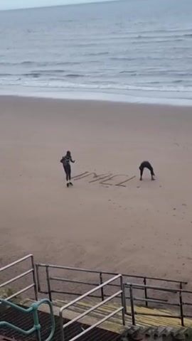 A person is writing in the sand on a beach. Another person is standing nearby and looking at them. The person writing in the sand appears to be drawing letters or symbols. The beach is sandy and there is a body of water in the background. There are some stairs and railings in the foreground. The sky is overcast.