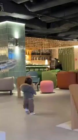 A toddler wearing a gray jacket and brown pants stands on a smooth, light-colored floor in what appears to be a modern lounge or waiting area. The toddler is holding two drumsticks and clapping them together. In the background is a bar area with illuminated shelves containing bottles. A variety of colorful seating, including chairs, ottomans, and couches are arranged around the toddler. A decorative column is covered in green and white tile. The ceiling above has exposed ductwork and lighting. A stroller is visible to the left of the toddler.