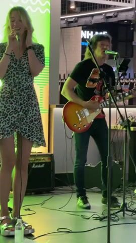 A male guitarist wearing a black t-shirt and jeans plays a cherry sunburst electric guitar and sings into a microphone. He has long, dark hair. A female singer in a leopard print dress stands to his left and also sings into a microphone. They are performing on a stage with a green and yellow backdrop. A Marshall amplifier is visible behind the female singer. The background shows a building with lights and windows. Cables and audio equipment are scattered on the stage floor.