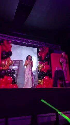 A person in a sparkly silver dress is on a stage singing into a microphone. They're standing in front of a large screen that shows a city skyline. There are red, gold, and black balloons on either side of the screen. The person raises their hands and moves around on the stage while singing. The stage is decorated with red fabric and there are chairs in front of the stage.