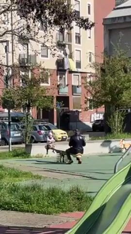 A small child in a light pink jacket rides a seesaw with an adult figure in a black jacket and brown pants, in an outdoor playground with a green surface. The playground includes a green slide in the foreground. In the background are multi-story residential buildings with various colors and architectural styles, as well as parked cars and greenery. The day appears bright with sunlight casting shadows in the playground.