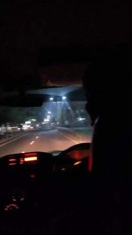 The video shows the interior of a car driving at night on a road with streetlights. The dashboard lights are visible, and a person's silhouette can be seen in the passenger seat. The road is lined with trees and buildings, and there are other cars on the road. The camera is pointed forward, giving a view of the road ahead.