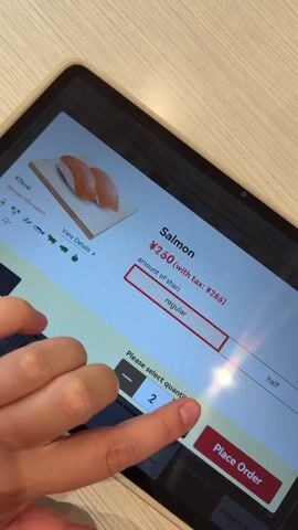 The video shows a hand interacting with a tablet screen. The screen displays a menu for ordering food, specifically sushi. The first frame shows a close-up of the tablet screen with an image of salmon sushi and options to select the amount of shari and quantity. The hand is seen touching the screen, seemingly to adjust the quantity or select an option. The subsequent frames show the screen updating as the user interacts with it. The price changes from ¥260 to ¥520, and the phrase "Thank you for your order" appears. The final frame shows a broader view of the tablet screen, displaying a variety of sushi options, including different types of salmon sushi, with prices listed for each. The overall context is of a person ordering food through a digital kiosk or tablet interface.