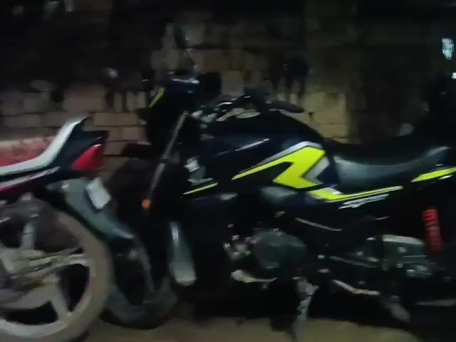 The video shows two motorcycles parked side-by-side in what appears to be a dimly lit indoor space, possibly a garage or shed. The motorcycle on the left is mostly visible from the rear, showcasing its black body, red taillight, and silver wheel spokes. The motorcycle on the right is a black sportbike with yellow and white accents on its body and a prominent front headlight. The background consists of a brick wall and some indistinct objects, suggesting a rustic or utilitarian setting. There are no people or animals visible in the video, and no actions or interactions are taking place.