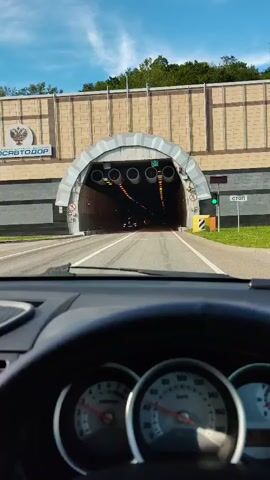 The video captures a first-person view from inside a car as it approaches and enters a tunnel. The dashboard and steering wheel of the car are prominent in the foreground.

**Scene and Setting:**
The scene begins outside the tunnel, under a bright blue sky, indicating daytime. The tunnel entrance is a large, gray, arched structure with a beige, rectangular building above it. Several large, cylindrical ventilation fans are suspended from the tunnel ceiling just inside the entrance. A green traffic light is visible to the right of the tunnel opening. As the vehicle enters, the tunnel interior becomes dark, lit by numerous square, warm-toned yellow lights arranged in rows along the ceiling and upper walls. The road inside is asphalt with clear white lane markings.

**Activity:**
The primary activity is driving a car into and through a tunnel.

**Subjects and Objects:**
- **Vehicle Interior:** A dashboard with two main circular gauges (speedometer and tachometer) and a steering wheel are visible.
- **Tunnel Features:** The tunnel entrance, interior walls, ceiling lights, and ventilation fans.
- **Other Vehicles:** A yellow taxi cab is briefly seen inside the tunnel, followed by a white car further ahead.

**Notable Details:**
- **Time of Day:** Sunny daylight outside the tunnel; artificial illumination inside.
- **Location Cues:** The architecture suggests a modern road infrastructure.
- **Speed:** The speedometer shows the vehicle entering the tunnel at around 60 km/h, increasing slightly to about 70 km/h within the tunnel.

**Visible Text:**
- On a sign above the tunnel entrance, partial text "ОДОРОГ" is visible, likely part of a Russian word related to roads.
- To the right of the tunnel entrance, a sign partially shows "CTO," possibly part of "СТОП" (Stop) or another word.