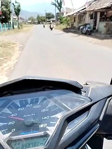 A first-person perspective video shows a rider on a motorcycle traveling down a paved road. The road is lined with buildings, some appearing to be small shops with corrugated roofs, and vegetation, including tall grasses and palm trees. Several motorcycles are visible on the road, with one in front of the rider and another further ahead. Red and white flags are displayed on poles near the buildings. In the background, mountains are visible under a clear blue sky. The rider's hand and a rearview mirror are visible in the foreground. The video captures the movement of the motorcycle as it navigates the road.