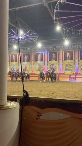 The video shows a horse arena with a sand floor. In the background, there is a stage with classical-style columns and paintings of figures. There are also decorative lights hanging from the ceiling, some of which are purple and appear to be part of a circular structure. In the arena, several horses are present, some with riders. One rider, dressed in blue, is seen galloping on a horse, holding a whip. Another horse and rider are moving towards the right side of the frame, near a colorful, star-shaped target. The scene appears to be a performance or show in a circus or similar venue.