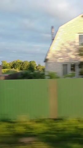 The video shows a blurred, side-view perspective of a rural landscape, likely from a moving vehicle. The foreground is dominated by a tall, light-colored fence. Beyond the fence, there are several buildings, including what appears to be a partially constructed structure with arched openings, a gray industrial-looking building with small windows, and residential houses. Green trees and vegetation are visible throughout the scene, with a blue sky and some clouds overhead. The motion blur suggests the camera is moving at a moderate speed. There are no people or clear interactions visible.