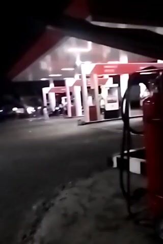 The video captures a nighttime scene at a gas station. The gas station features a canopy with red accents and fluorescent lighting. Several vehicles, including what appears to be a truck and multiple motorcycles, are present. People are standing around, some near the vehicles and others near the gas station buildings. One individual is seen on a motorcycle. The ground is paved and appears somewhat rough. The overall atmosphere is dark, with the gas station lights providing illumination. There are also some structures visible in the background.