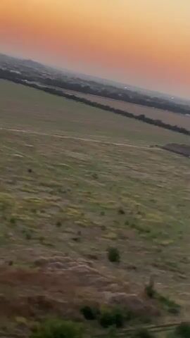 The video captures a panoramic view of a rural landscape during sunset. The sky transitions from a warm orange hue near the horizon to a pale yellow higher up. Below the sky, a vast expanse of green fields stretches out, with some areas appearing dry and brown. A line of trees marks the border between the fields and a distant town, which is visible as a cluster of buildings and lights. The camera pans slowly, revealing subtle details such as mounds of dirt or debris in one of the fields and what appears to be a white building or structure on the far right edge of the frame. The overall scene conveys a sense of calm and spaciousness, with the natural light of the setting sun casting a soft glow over the landscape.