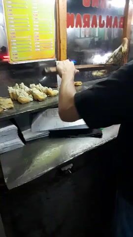 A person wearing a black t-shirt and blue jeans stands behind a metal counter. The person is holding a pair of wooden tongs and appears to be preparing food. On the counter, there are several plates of fried food items, possibly plantains or fritters. To the left of the counter, a yellow menu board is visible. Behind the counter, a glass window displays a red sign with white text, which reads "UNGGU BANGARAN". The environment appears to be an outdoor food stall at night, with artificial lighting illuminating the scene.