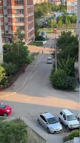A video pans upward over a street scene. In the foreground, two cars are parked. A grey sedan is in the left foreground, and a white SUV is in the right foreground. A paved path runs along the left side of the frame, bordered by a low metal fence and some vegetation. A person in a pink outfit walks along this path. Further up the path, two people in light-colored clothing walk away from the camera. A white SUV drives away from the camera on the paved street. To the right of the street, there are many trees and a gray utility pole with wires. Further down the street, a red and white vehicle, possibly a bus or truck, is visible. The background shows several multi-story buildings, some with balconies, and more trees and streetlights. The sky is a pale blue.