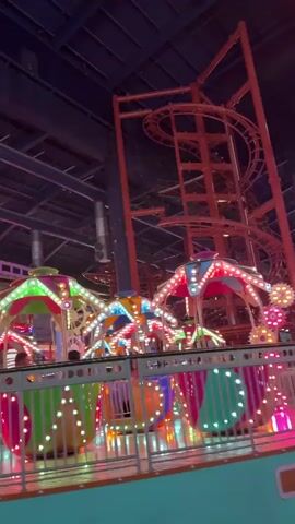 The video shows an indoor amusement park with multiple brightly lit rides and a roller coaster. The foreground features a spinning ride with colorful, striped gondolas. Above, a small roller coaster train is seen moving along a track. The overall lighting is dim, but the rides themselves are illuminated with vibrant colors. The video is shot from a moving perspective, as if the camera is on another ride passing by.