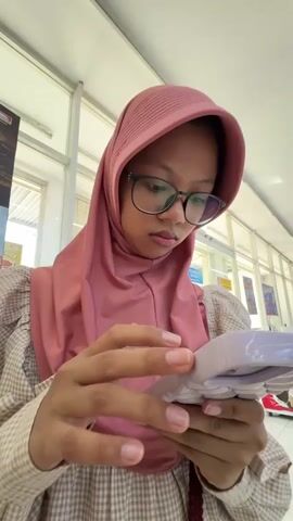 A medium shot captures a young person wearing a pink hijab and glasses, focused on a device held in their hands. The individual's face is partially obscured by the glasses, and their expression appears serious or contemplative. The person is wearing a beige and brown plaid top with puffed sleeves. Their hands are in focus as they manipulate the device. The background suggests an indoor environment, possibly a public space with bright lighting and architectural elements like ceiling panels and what appear to be windows or partitions. There are hints of posters or displays in the far background, suggesting a retail or communal area. The overall scene is static, with no discernible movement beyond the manipulation of the device.