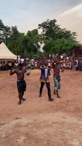 In an outdoor setting with reddish-brown dirt, three men perform in the foreground while a large crowd observes in the background. The scene is framed by lush green trees under an overcast sky, suggesting late afternoon or early evening.

The man on the left is shirtless, wearing patterned shorts and sandals, and holds a light-colored cloth or object in his left hand while flexing his right arm. The man in the center wears a dark blue patterned shirt, black pants, and sandals, holding a stick or staff. The third man, on the right, is shirtless, dressed in patterned shorts and sandals, and also holds a stick or staff. They appear to be engaged in a synchronized dance or ritualistic performance.

Behind them, a dense crowd of people, including many children, watches intently from a slight distance. A white tent is visible on the far left of the background. The overall atmosphere suggests a communal gathering or celebration in Achara, Nigeria.