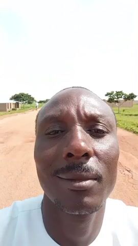 A close-up selfie video shows a man with dark skin, a mustache, and a shaved head. He is wearing a white collared shirt. The man is outdoors on a dirt road. In the background, there are a few cars, some low buildings, and green vegetation under a bright sky with some clouds. The camera appears to be moving.