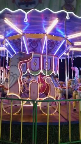Here is a description of the video:

The video shows a carousel operating at night. The carousel is lit up with colorful lights. The horses are painted in various colors and are moving up and down as the carousel spins. There is a decorative carriage in front of the horses. A green and yellow fence is around the carousel.  A person is briefly seen walking past the fence.