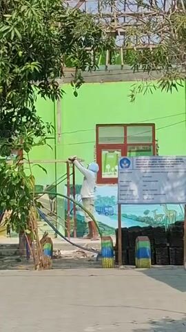 A person is seen on a structure, possibly constructing or repairing it. The person is wearing a light-colored shirt and pants, and a head covering. They are holding a pole or pipe and appear to be attaching it to the structure. The background shows a bright green building with a red-trimmed window. There is also a mural or painting on the building depicting animals and a truck. Signs are posted next to the building, one with the Indonesian national emblem. Trees with green leaves and what appear to be mangoes are visible. The ground is paved and appears to be an outdoor area, possibly a playground or schoolyard.
