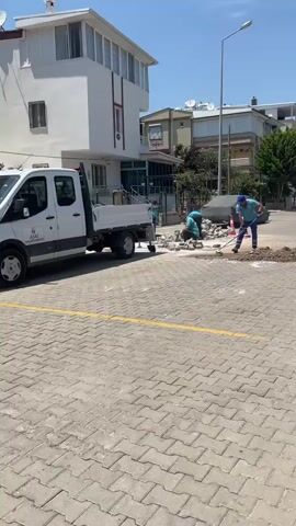 A group of construction workers are seen working on a street. One worker is pushing a broom, while another is kneeling and appears to be laying down pavers. A third worker is also kneeling and is surrounded by stones. A white truck is parked on the left side of the frame, and a car covered with a tarp is visible in the background. The scene is taking place in an outdoor urban environment on a sunny day.