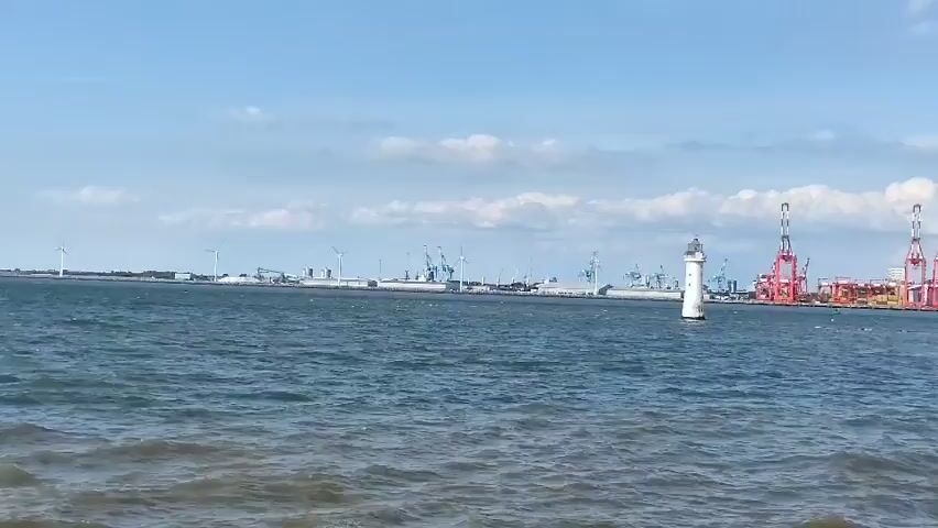 The video shows a wide shot of a harbor on a sunny day with a blue sky and scattered clouds. In the foreground, choppy blue water laps at a sandy shore. In the middle ground, a white lighthouse stands in the water. Further in the distance, a large industrial port is visible, featuring numerous red shipping cranes, buildings, and wind turbines. Several people are visible on the beach in the far right of the frame.