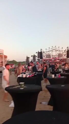 The video shows an outdoor event on a sandy beach with a stage and DJ setup in the background. Many people are gathered around black cocktail tables, some standing, some socializing. The sky is clear and the lighting suggests it is late afternoon or early evening. The atmosphere appears festive with people dressed casually. The stage features a large sound system and lighting equipment, indicating a musical performance is or will be taking place.