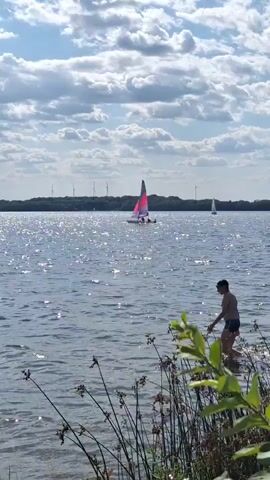 A person is walking in the water at the edge of a lake. There is a sailboat in the distance, as well as wind turbines on the horizon. The water is sparkling in the sunlight, and there are clouds in the sky. The person is wearing swim trunks and appears to be walking towards the deeper water.
