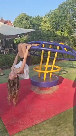 In a park setting, likely Groenlo, Netherlands, a young girl is captured in a series of frames performing a handstand on a children's merry-go-round. The merry-go-round itself is a vibrant play structure, featuring a blue frame with yellow circular supports, all mounted on a purple circular base. It sits on a red, textured play surface.

The girl, with long brown hair, is dressed in a white top and dark shorts. In the first frame, she is mid-flip, with her body angled downwards, hair cascading. As the merry-go-round rotates, she transitions into a handstand, with her legs extended upwards. A second child, with blonde hair and wearing a white top and patterned trousers, is visible in the background of one frame, observing or possibly waiting to play.

The scene appears to be set outdoors during the daytime, with clear blue skies and bright sunlight casting shadows, suggesting fair weather. Lush green trees and foliage surround the play area, with a hint of a building with a red roof and a canopy structure visible in the distance, possibly an outdoor cafe or seating area. The overall mood seems playful and energetic, with the child clearly enjoying her acrobatic display on the playground equipment. No visible text is present in the media.