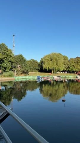 A vertical video pans right across a calm, dark body of water. On the left bank, a green fence runs along the edge, backed by lush green trees and a tall white communication tower. Several boats are moored along the bank, including a blue motorboat, a white speedboat with red trim, and other smaller vessels. A large, leafy green tree with drooping branches stands out among the others. On the right side of the water, more boats are docked, some with white covers. Small orange buoys are visible floating in the water, connected by lines. Further in the distance, more boats and buildings line the waterway, with trees and structures visible on the far bank under a clear blue sky. The reflections of the trees and boats are visible on the water's surface.