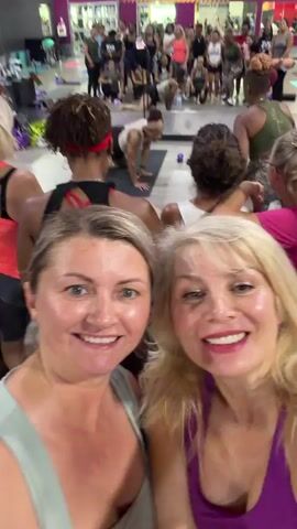 The video begins with a selfie-style shot of two women smiling at the camera, appearing to be in a fitness class. In the background, other participants are visible, some sitting on the floor and others standing. A large purple banner with white text that reads "FAMILY CLASS-IFIED AREA" is prominently displayed on the wall behind them. As the video progresses, the camera pans slightly to show more of the class in session, with people in athletic wear and various equipment like mats and small weights. The lighting suggests an indoor setting, possibly a gym or studio. The overall mood appears energetic and positive.